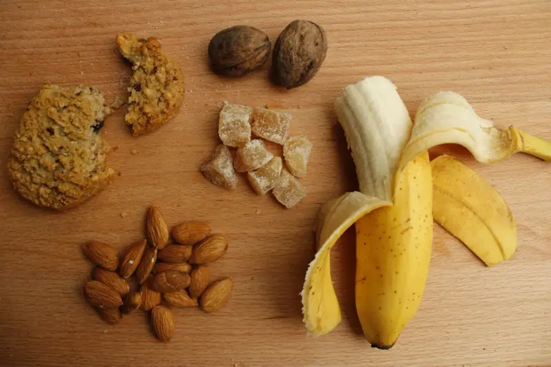 Na drewnianym blacie leżą orzechy, migdały, kawałek ciastka, kandyzowany imbir i banan. To świetne węglowodany złożone produkty.