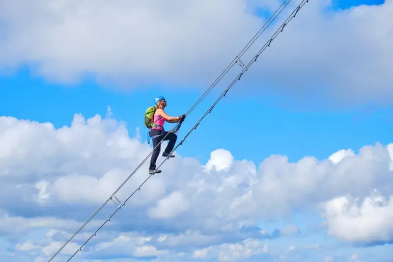 Frau klettert auf dem Donnerkogel Klettersteig über eine Hängebrücke, umgeben von blauem Himmel und Wolken.