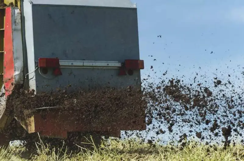 Rozrzutnik rozsypuje komunalne osady ściekowe na polu. Materiał jest wyrzucany w powietrze, tworząc brązową chmurę.