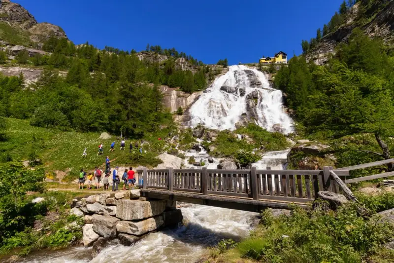 Cascata imponente con escursionisti su un sentiero, un ponte di legno attraversa un torrente. Un esempio di cosa vedere in Piemonte per gli amanti della natura.