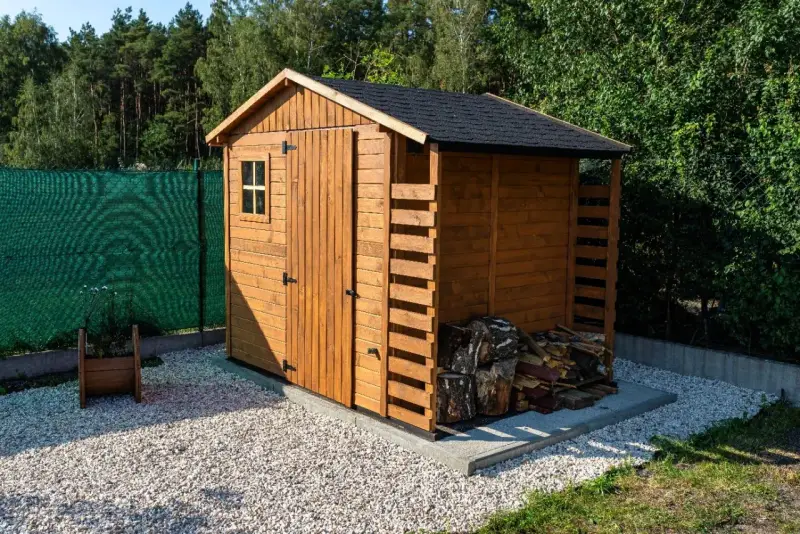 Drewniany domek na narzędzia ogrodowe, z małym okienkiem i drzwiami, obok stos drewna.