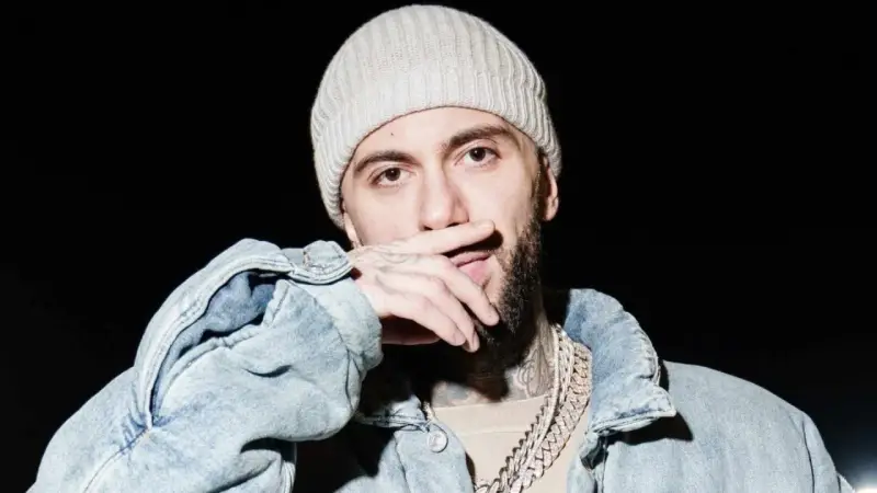Malik Montana w czapce i jeansowej kurtce, z dłonią na ustach, gotowy na koncert.