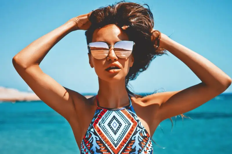 Kobieta w okularach przeciwsłonecznych na plaży, z rękami w gorze. Jej skóra ma piękny, złocisty odcień, jakby świadczył o wspaniałych beta karoten efektach.