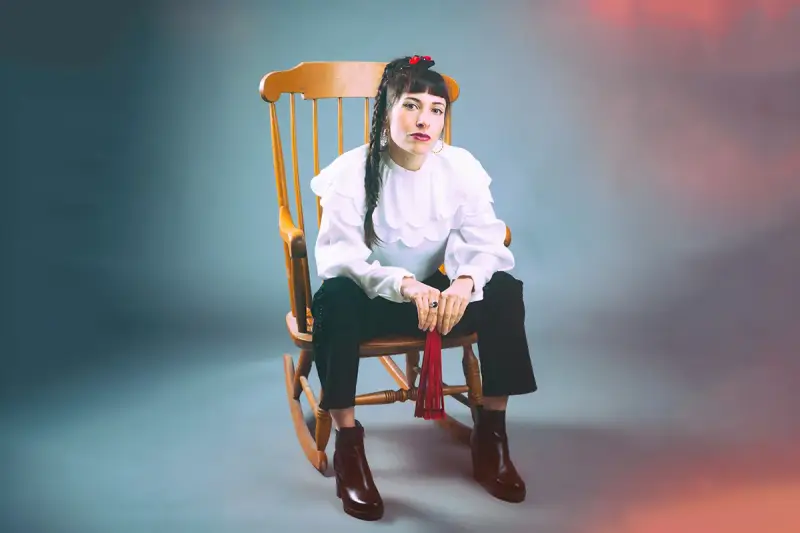 Scout Niblett, assise sur un fauteuil à bascule, porte une blouse blanche à volants et un pantalon noir. Elle tient un pompon rouge.