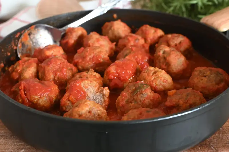 Un tegame pieno di polpette al sugo, pronte per essere gustate. Un cucchiaio d'argento è appoggiato sul bordo, invitando a servirle.