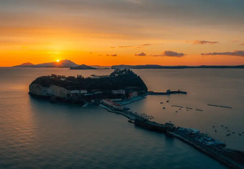 Tramonto infuocato sul golfo di Napoli, con una suggestiva isoletta che emerge dal mare calmo, illuminata dagli ultimi raggi dorati.