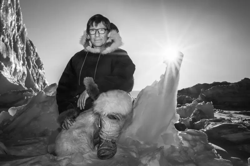 Ein Mann in traditioneller Inuit-Kleidung sitzt in eisiger Landschaft. Die Sonne scheint hell.