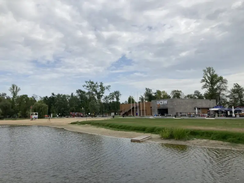 Spokojne jezioro małe żnińskie z piaszczystą plażą, budynkiem CSW i zielenią w tle.