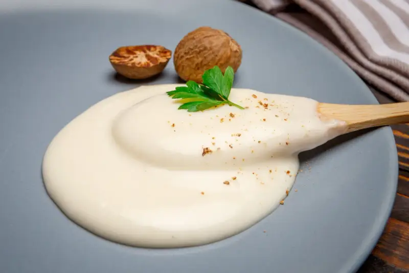 Krämig bechamelsås på en tallrik med muskotnötter och persilja. Ett enkelt bechamelsås recept för alla.