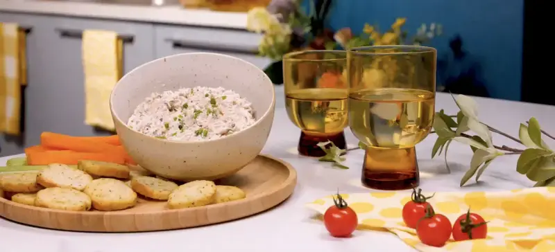 Un bol de rillettes de thon crémeuses, garni de ciboulette, servi avec des crackers, des bâtonnets de carottes et de céleri. Deux verres de vin blanc complètent cette scène gourmande.