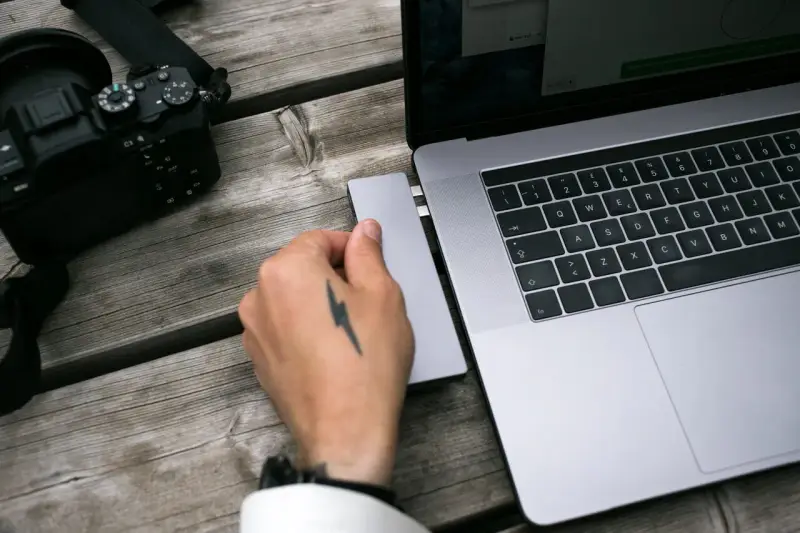 Dłoń z tatuażem pioruna podłącza adapter do laptopa, obok leży aparat. Idealny zestaw, jaki laptop do obróbki zdjęć?