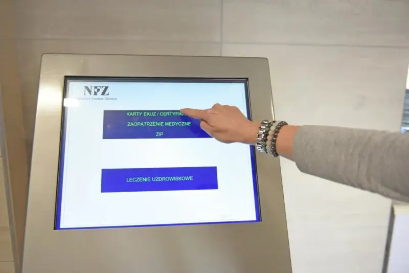 Dłoń dotyka ekranu automatu NFZ, wybierając opcję "Karty EKUZ / Certyfikaty".