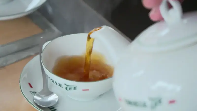 Tee mit Rum norddeutsch: Eine weiße Kanne gießt heißen Tee in eine Tasse. Ein Löffel liegt auf der Untertasse.