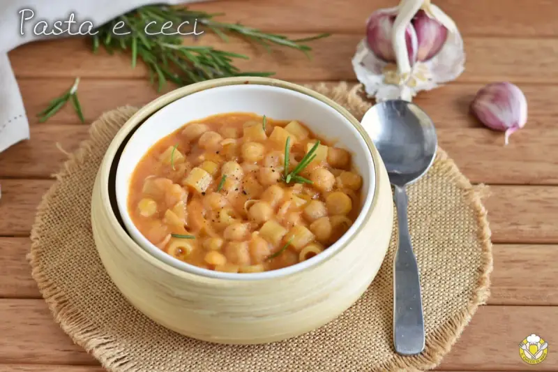 Un piatto fumante di pasta e ceci, guarnito con rosmarino fresco, su un tavolo di legno. Accanto, spicchi d'aglio e un cucchiaio.