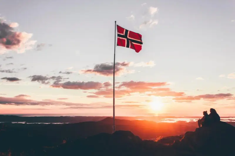 Zachód słońca w Norwegii. Dwie osoby siedzą na skale, patrząc na krajobraz. Flaga Norwegii powiewa na maszcie. Czy do Norwegii można lecieć na dowód osobisty?