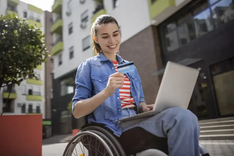 Uśmiechnięta kobieta na wózku inwalidzkim, korzystająca z laptopa i karty płatniczej, pokazuje, że stopnie niepełnosprawności nie ograniczają jej aktywności.
