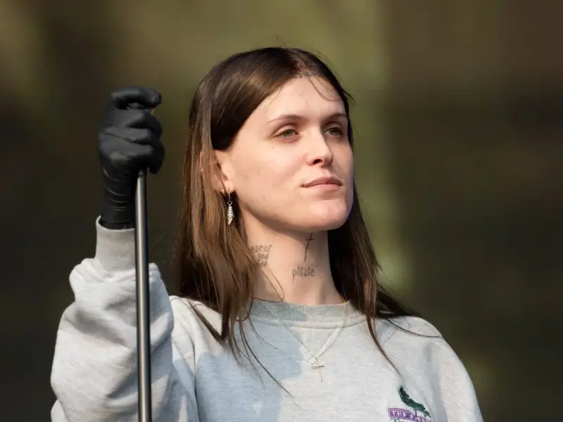 Ethel Cain, con guantes negros y un collar de cruz, mira hacia un lado con una expresión pensativa.