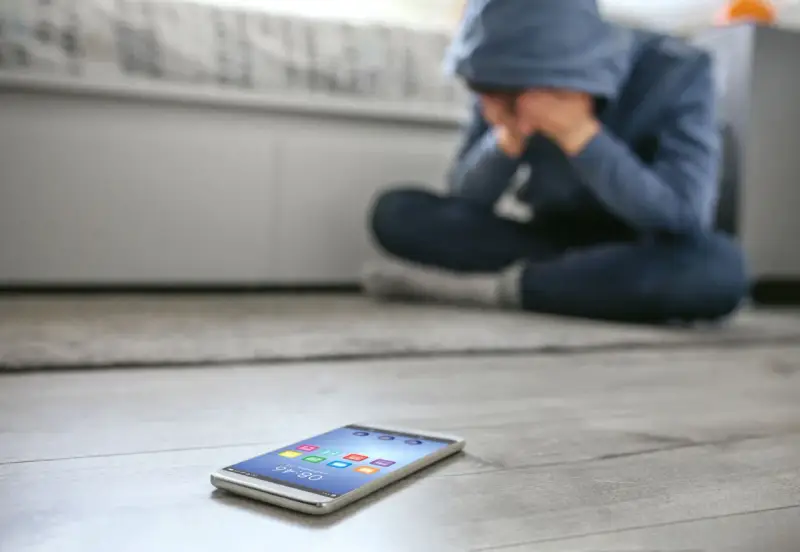 Osoba w kapturze siedzi na podłodze, ukrywając twarz w dłoniach. Obok leży telefon, symbolizujący izolację i dystymię.