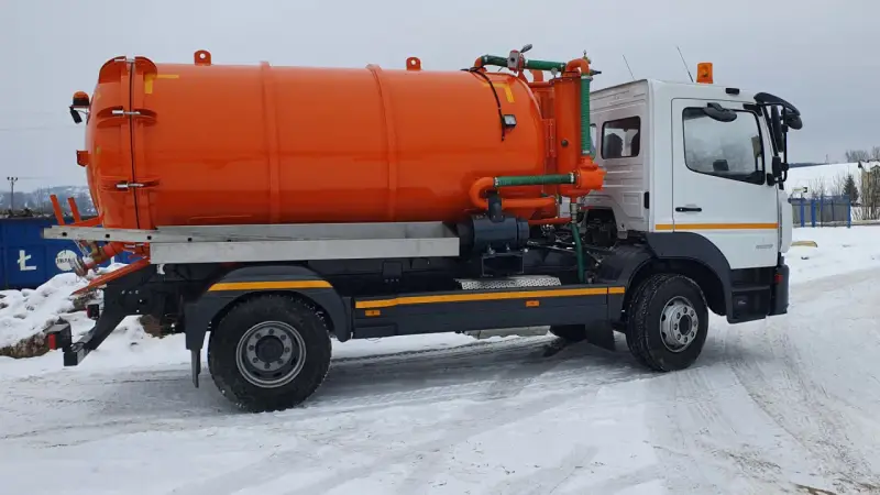 Pomarańczowy pojazd asenizacyjny z beczką i zielonymi wężami stoi na zaśnieżonej drodze.
