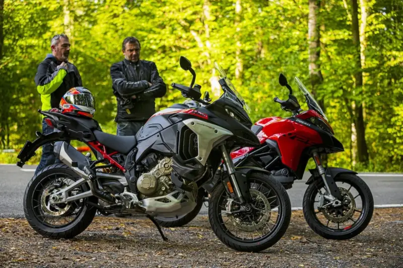 Dwóch motocyklistów z kaskiem na siedzeniu obok dwóch motocykli Ducati Multistrada, szary i czerwony, na tle leśnej drogi.