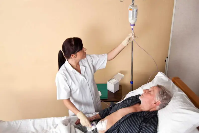 Pielęgniarka podłącza kroplówkę do pacjenta leżącego w łóżku. Widać rurkę od PEG.