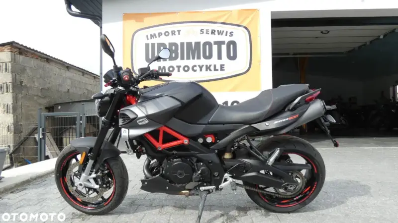 Czarny motocykl Aprilia Shiver 900 z czerwonym stelażem, zaparkowany przed salonem motocyklowym.