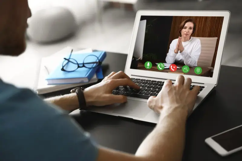 Mężczyzna prowadzi wirtualną superwizję przez laptopa. Na ekranie widać kobietę z dłońmi złożonymi w geście modlitwy.