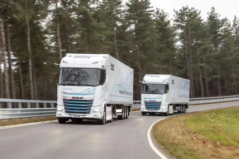 Dwa białe elektryczne DAF XG Plus z naczepami jadące po krętej drodze wśród drzew.