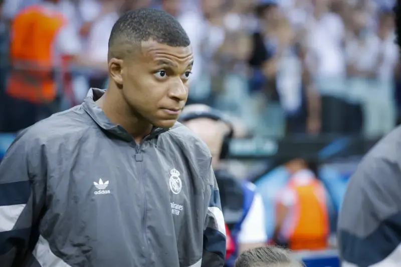 Kylian Mbappé w szarej kurtce z logo Realu Madryt, gotowy do gry.