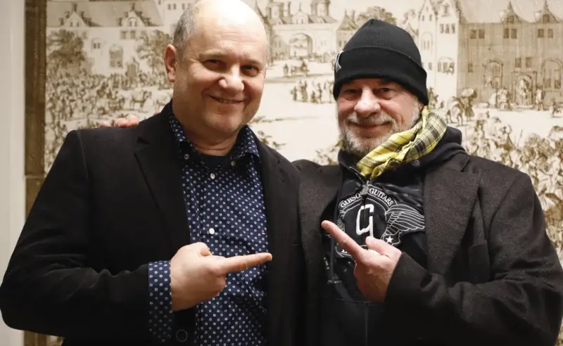 Deux hommes souriants, l'un avec une chemise à pois, l'autre avec un bonnet, se montrent du doigt. Un chanteur de confiance.