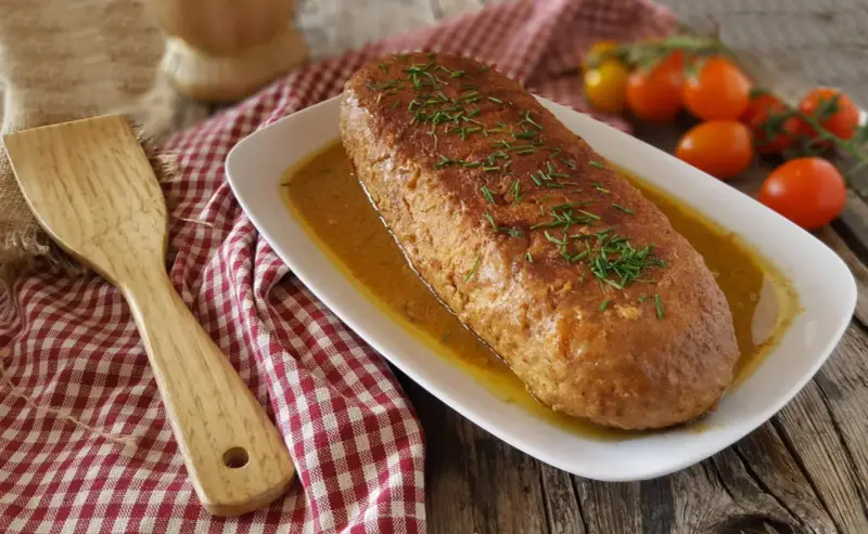 Polpettone in padella dorato, guarnito con erba cipollina, servito su un piatto bianco con salsa. Accanto, una spatola di legno e pomodorini.