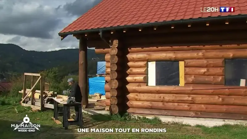 Une maison en rondins de bois en construction, avec un homme travaillant à proximité. Le toit est rouge et le paysage est montagneux.