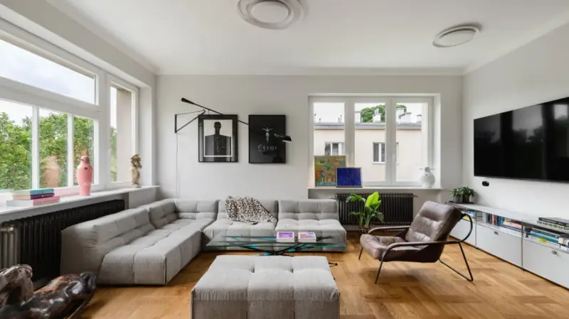 Nowoczesny salon z szarym narożnikiem, szklanym stolikiem i brązowym fotelem. Widać dzieła sztuki, rośliny i telewizor. To przykład udanego **retrofit** mieszkania.