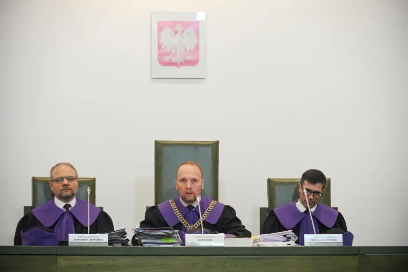 Trzech sędziów w togach podczas jawnego posiedzenia sądu. Na ścianie wisi godło Polski.