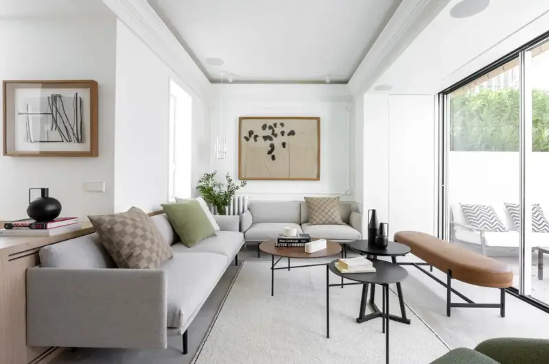 Salón moderno con sofás grises, mesas bajas negras y marrones, y un banco de cuero. Grandes ventanales conectan el interior con la terraza, creando un espacio fluido para unir terraza y salón.