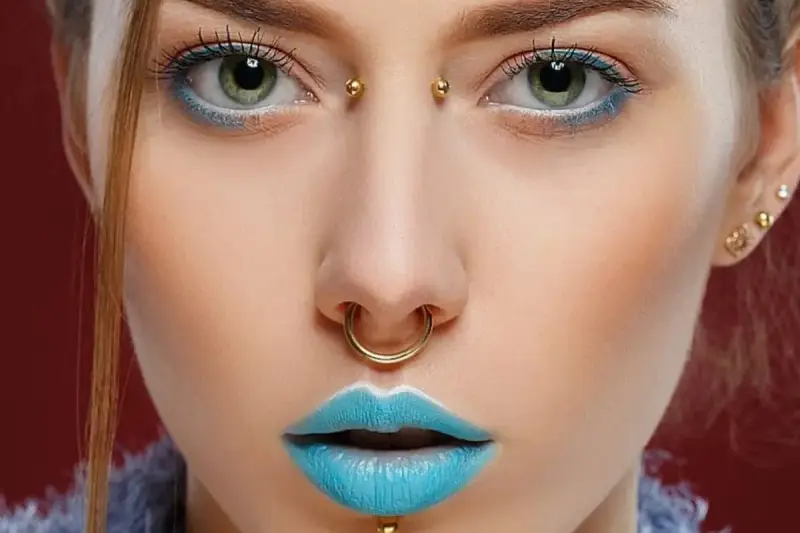 Kobieta z błękitnymi ustami i złotym septum. Jej oczy podkreślone są niebieskim cieniem, a między brwiami widoczny jest bridge piercing.