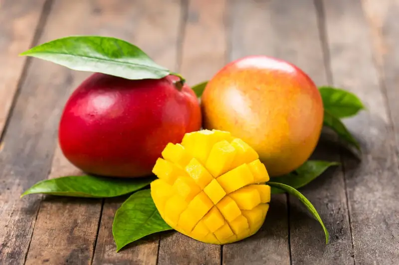 Dwa soczyste mango, jedno czerwone, drugie pomarańczowe, leżą na drewnianym blacie. Obok nich pokrojone w kostkę, złociste wnętrze owocu. To przypomina o tropikalnych krajach, skąd pochodzi mango.