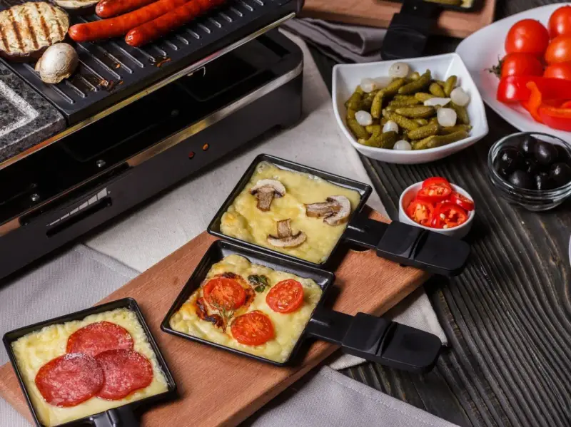 Raclette z pieczarkami i pomidorami, obok kiełbaski. Idealny ser do raclette rozpuszcza się w tych małych patelenkach.