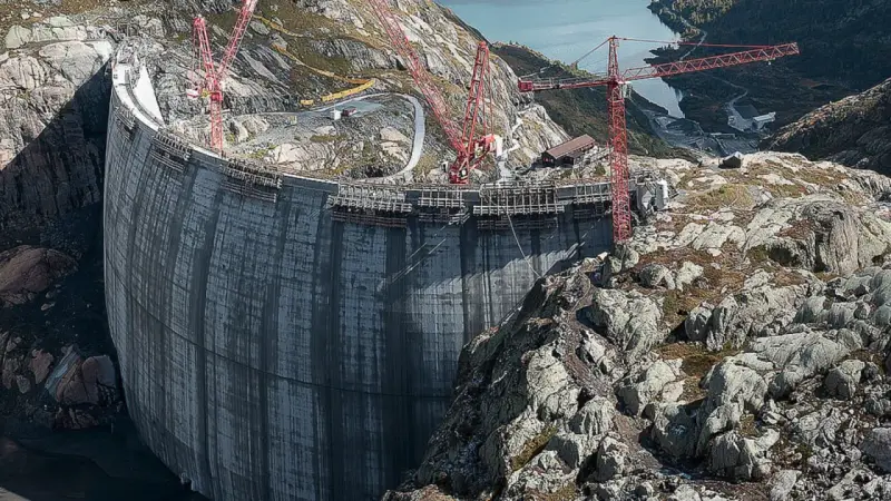 Bau einer gewaltigen Staumauer, die an Pumpspeicherkraftwerke in Deutschland erinnert. Kräne ragen in den Himmel, während die Betonkonstruktion wächst.