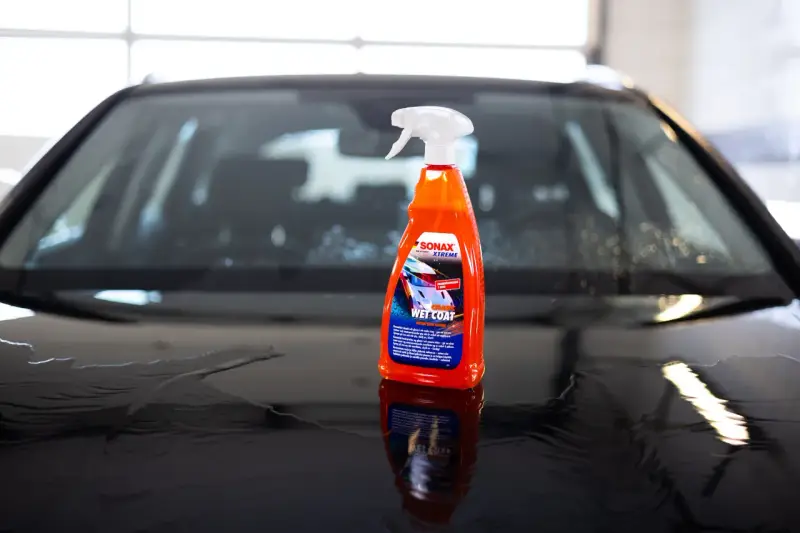 En flaska SONAX Xtreme Wet Coat står på en våt, mörk bilhuv.