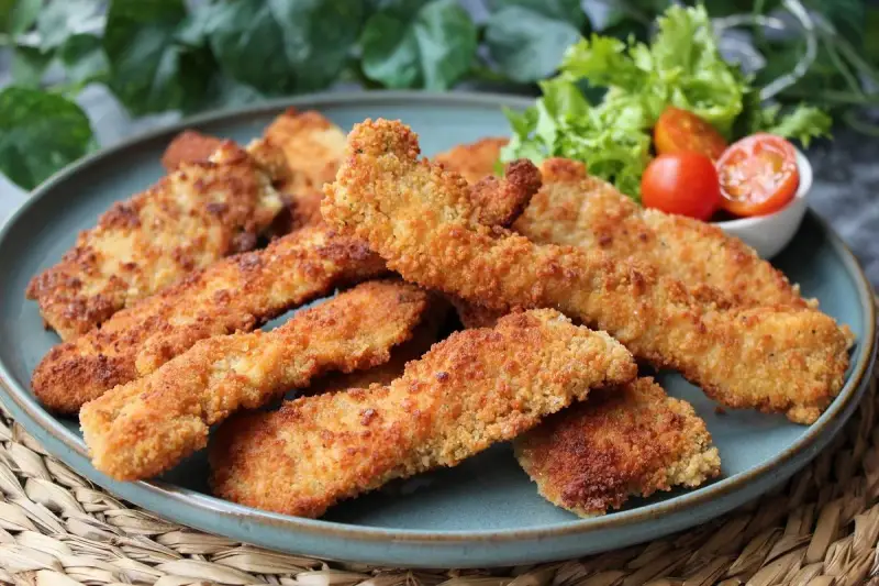 Crujientes fingers de pollo dorados en un plato azul, acompañados de lechuga y tomates cherry. ¡Perfectos para picar!