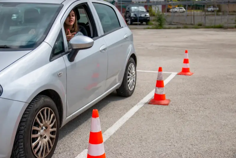 Młoda kobieta za kierownicą srebrnego samochodu ćwiczy jazdę na placu manewrowym. To może być jej droga do zdobycia prawa jazdy 17 lat.