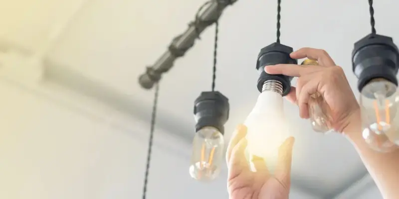 Manos cambian una bombilla LED por una incandescente en una lámpara colgante, actualizando la luz del hogar.