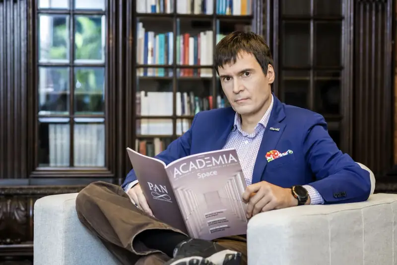 Mężczyzna w garniturze czyta magazyn "Academia Space" w bibliotece. W tle regały z książkami.