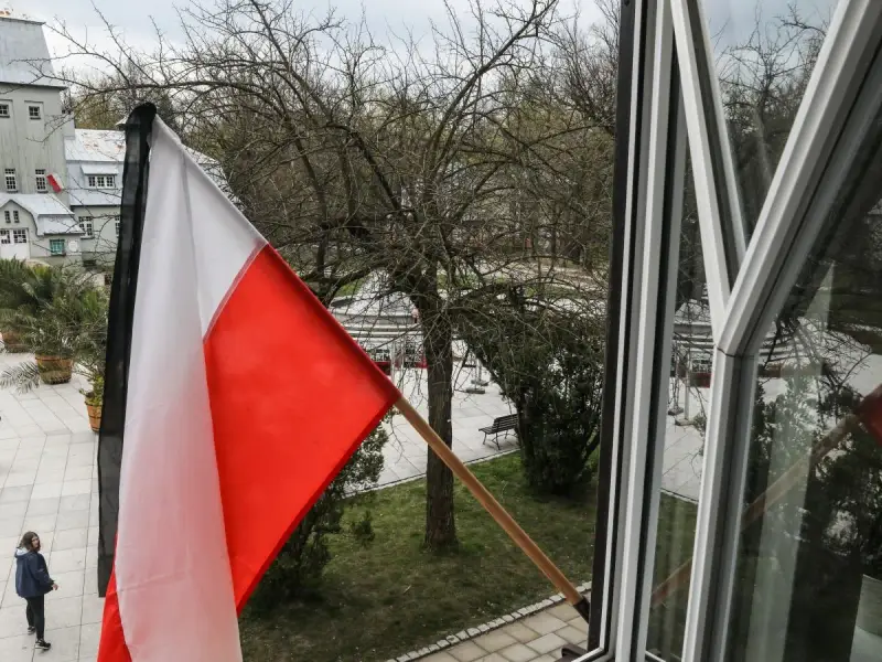 Flaga państwowa w biało-czerwonych barwach powiewa na wietrze. W tle widać drzewa i budynki.