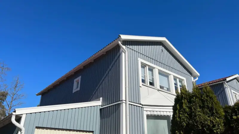 Blå himmel över ett gråblått trähus. Snart dags att måla fasad för att skydda det mot väder och vind.