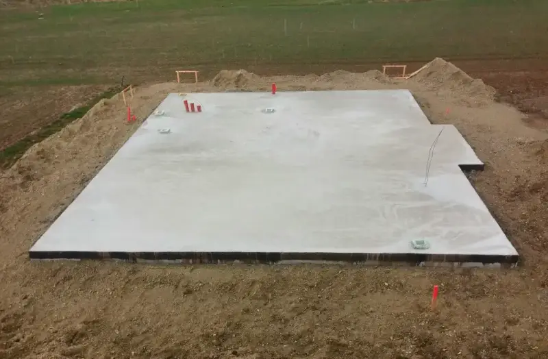 Płyta fundamentowa koszt budowy domu. Betonowa płyta fundamentowa z wystającymi rurami i elementami instalacji, otoczona ziemią i żwirem.