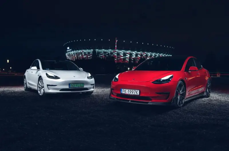 Dwa modele Tesla, biały i czerwony, zaparkowane nocą przed oświetlonym stadionem.