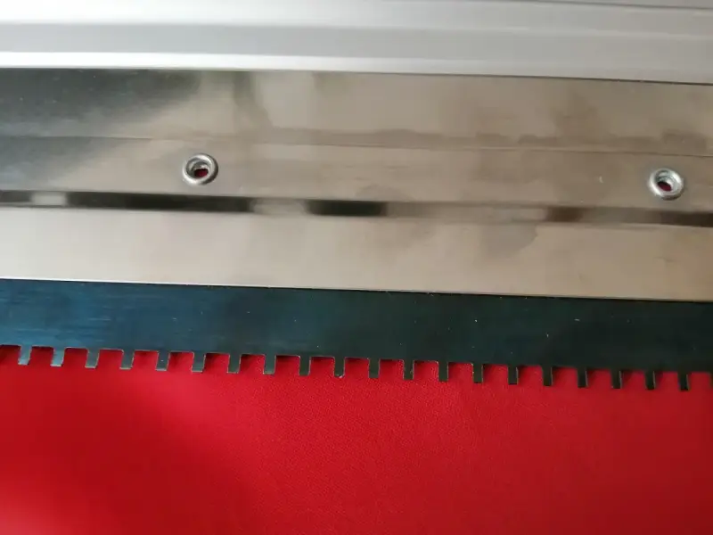 Metalowa rakla z ząbkowanym ostrzem, gotowa do pracy na czerwonym materiale.