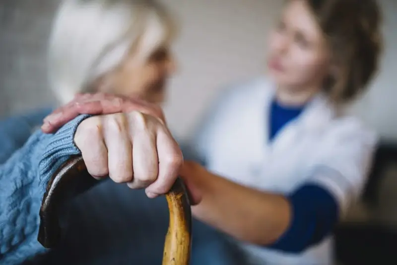 Opiekunka wspiera starszą panią, która trzyma laskę. Zastanawiasz się, ile kosztuje opiekunka z mops?